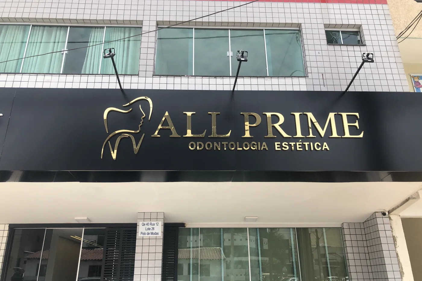 A fachada da All Prime vista de frente, com o letreiro dourado sobre a placa preta e a entrada de vidro embaixo.